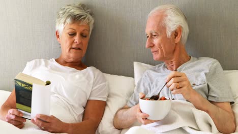 Senior-man-having-breakfast-while-woman-reading-a-book-on-bed