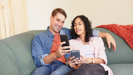 laughing together, young multiracial couple sitting on couch and using smartphones, at home