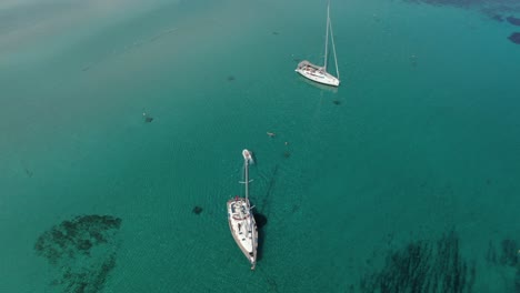 A-drone-shot-circling-two-sailing-boats-anchored-in-a-small-gulf-with-crystal-clear-blue-water