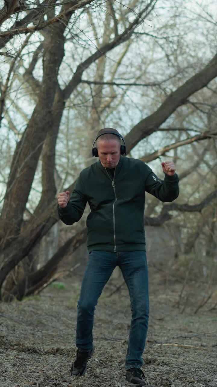 A man in a green jacket, headphones on, dances joyfully in a serene forest, he embodies freedom and happiness amidst the tranquil natural setting, with trees and leaves around him