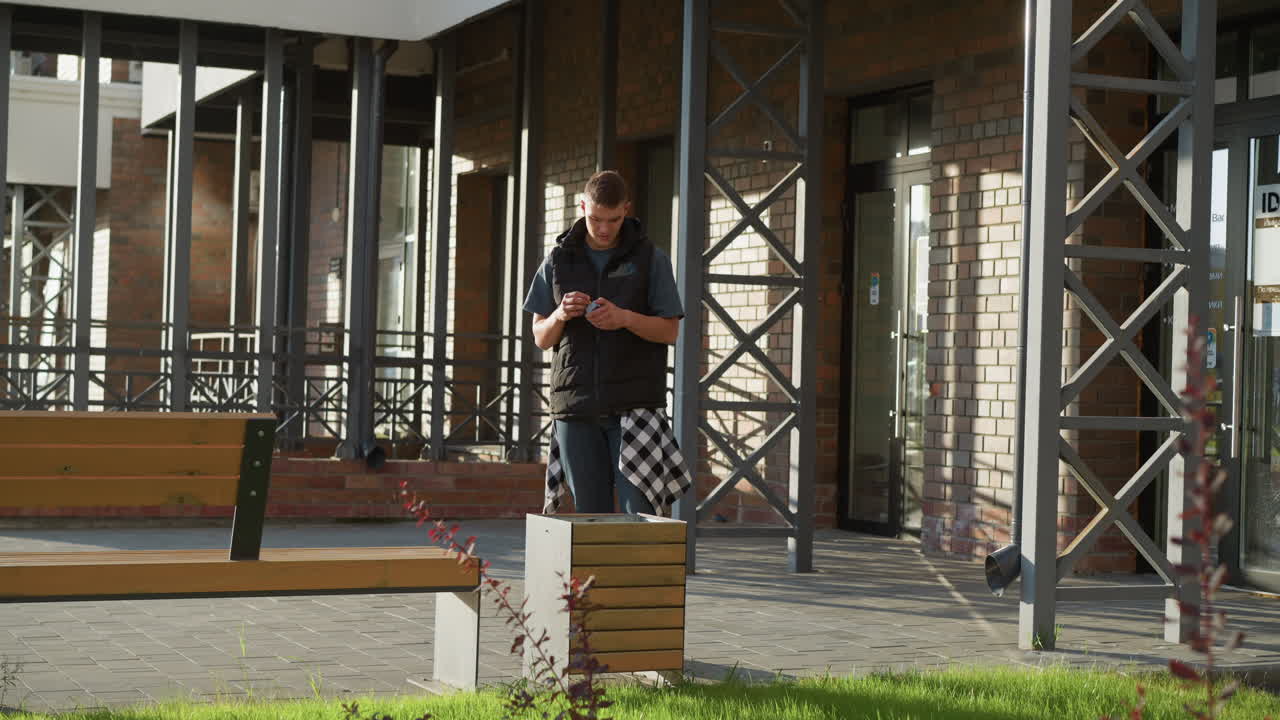 young man under canopy of brick facade pulls out cigarette inspects hesitates in bright daylight then tosses cigarette into waste bin by bench in urban courtyard