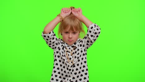 a young girl with blonde hair smiles happily as she poses in front of a green screen. she is wearing a white and black shirt with gold jewelry.