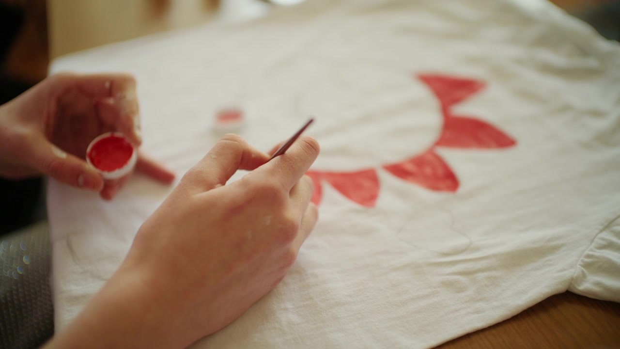 Primo piano di un artista che dipinge forme decorative rosse su una maglietta bianca usando pittura per tessuti