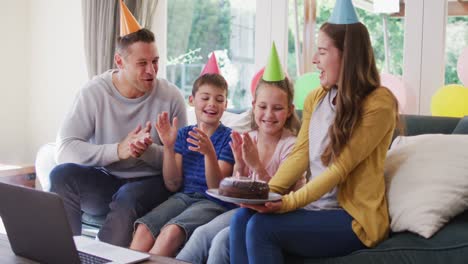 caucasian woman in party hat holding birthday cake while family clapping and celebrating birthday at