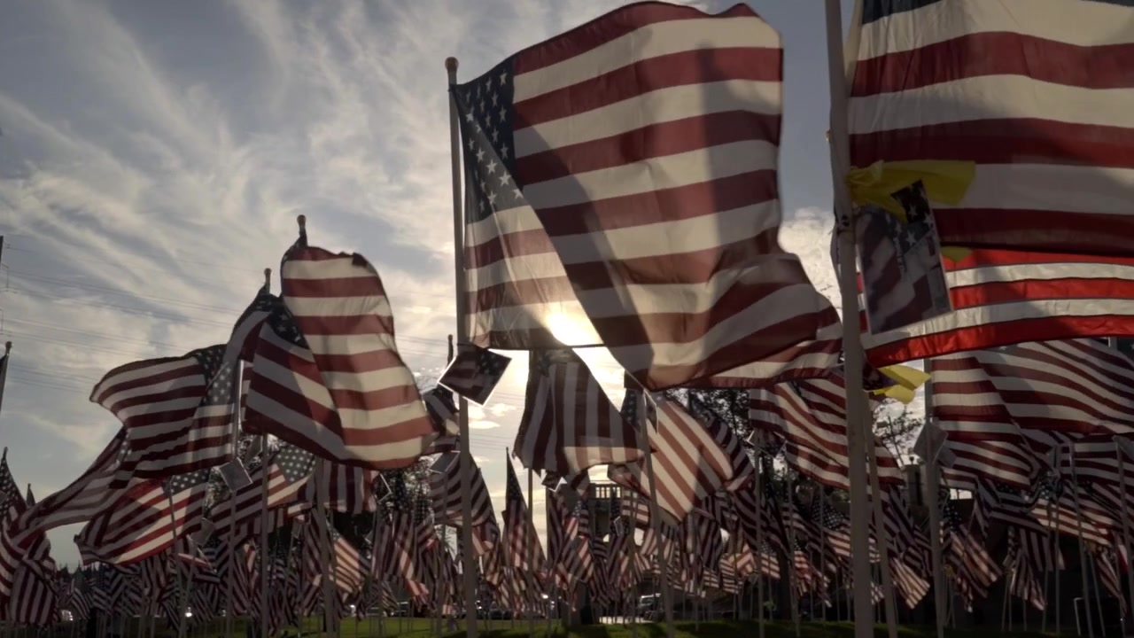 A slow zoom in on backlit American flags blowing in the wind at an outdoor memorial park. A patriotic display honoring Independence Day and national heroes with no people visible against a cloudy sky.