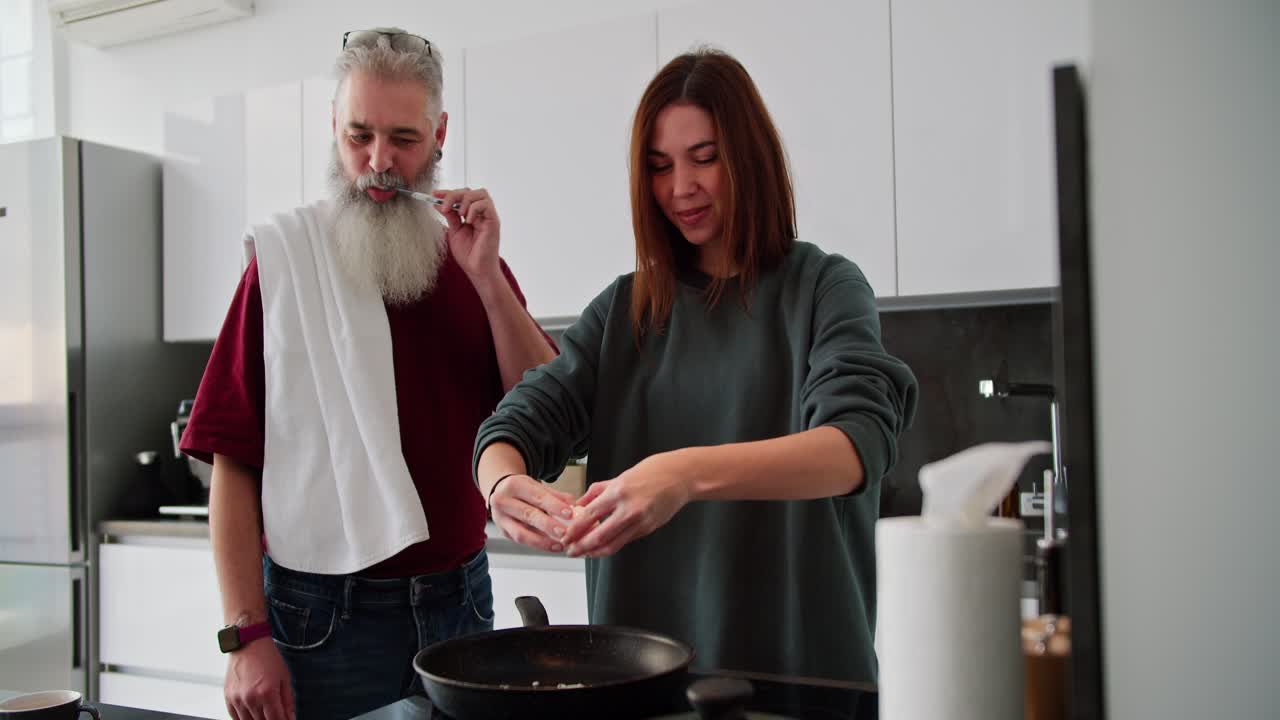Happy elderly man with gray hair and lush beard brushes his teeth with a towel on his shoulders while his adult brunette daughter in a green sweater makes breakfast in a modern kitchen