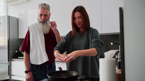 Happy-elderly-man-with-gray-hair-and-lush-beard-brushes-his-teeth-with-a-towel-on-his-shoulders-while-his-adult-brunette-daughter-in-a-green-sweater-makes-breakfast-in-a-modern-kitchen