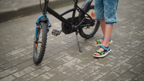 leg view of a young child parking a black bicycle on a paved path, the child carefully holds the handlebar while adjusting the bike s stand