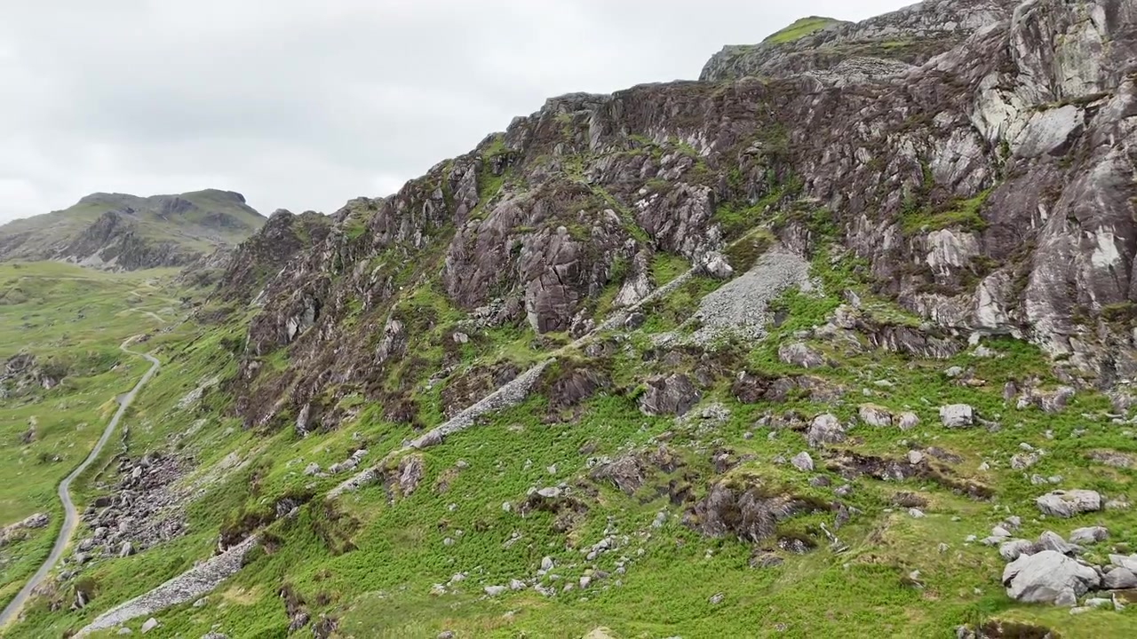 Jævn droneflyvning langs barske klippefyldte bjergskrænter i Snowdonia Nationalpark, Wales. Dramatisk naturlandskab med blotlagte klippeformationer og grøn vegetation fanget i filmisk 4K.