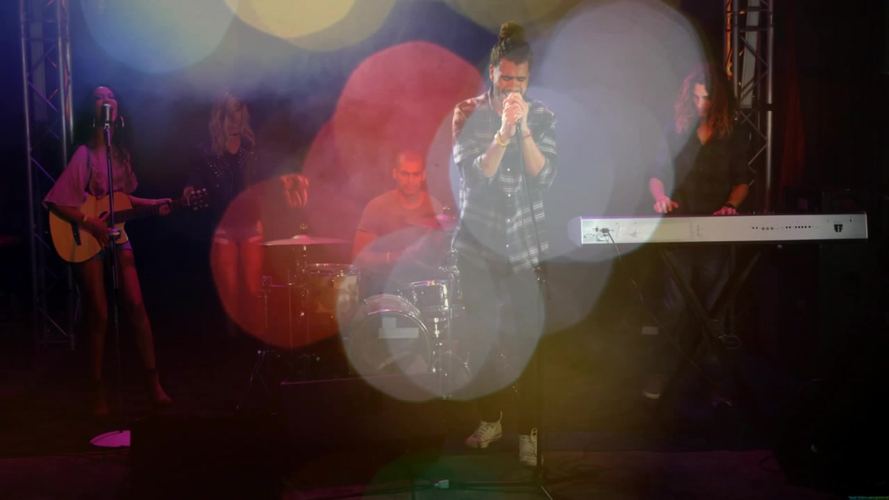 Vocalista segurando o microfone, cantando em palco com pouca luz, orbes de bokeh se deslocando, mostrando a banda para um show de música.