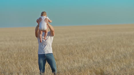 padre joven amoroso rodea a una hija bebé en el campo. padre cariñoso mira al bebé, él es feliz. hermoso paisaje rural, cielo azul. 4k, prores