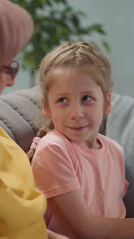 Adorable girl reads story and looks at senior woman seeking approval smiling in living room. Home education and leisure. Granny takes care of child