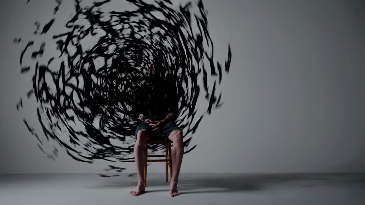 Barefoot man sitting on a chair against a white wall with swirling, dark, abstract shapes surrounding his head, creating a surreal and chaotic portrait