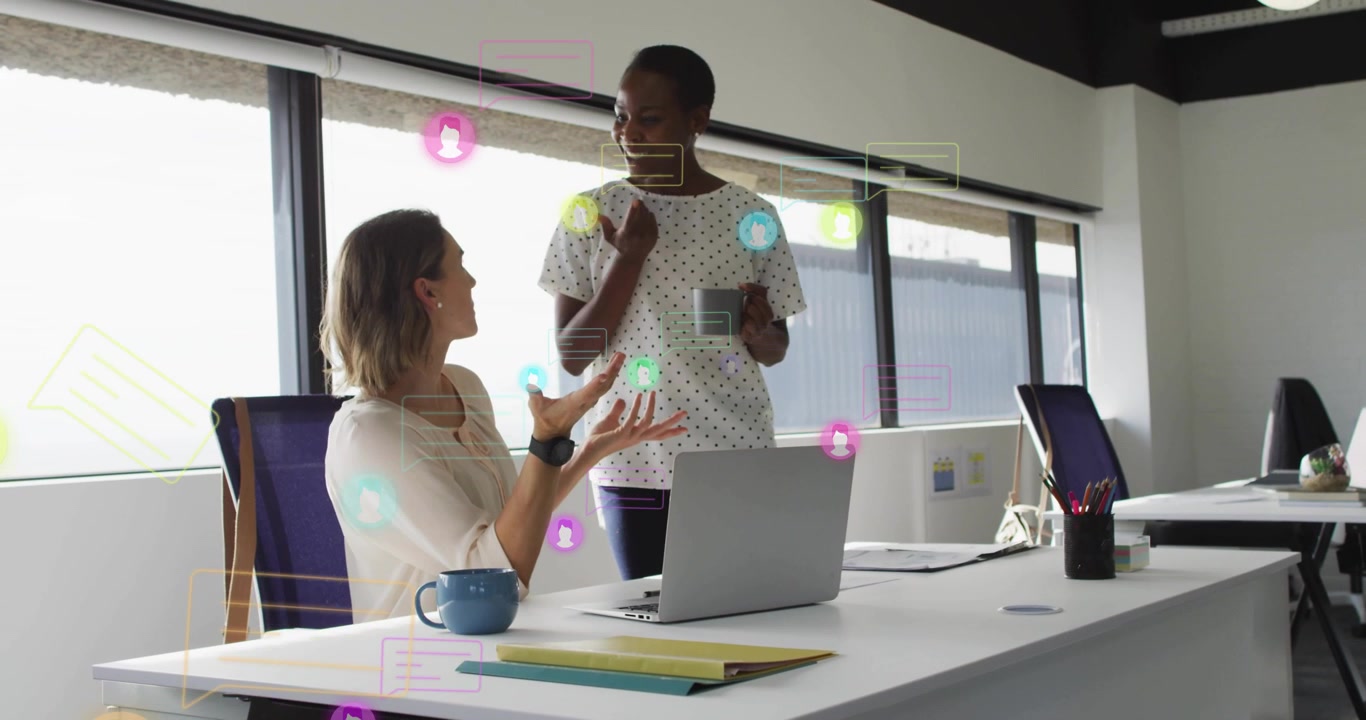 Seated woman gesturing at laptop causing chat alerts over desk while office peer nodding, working
