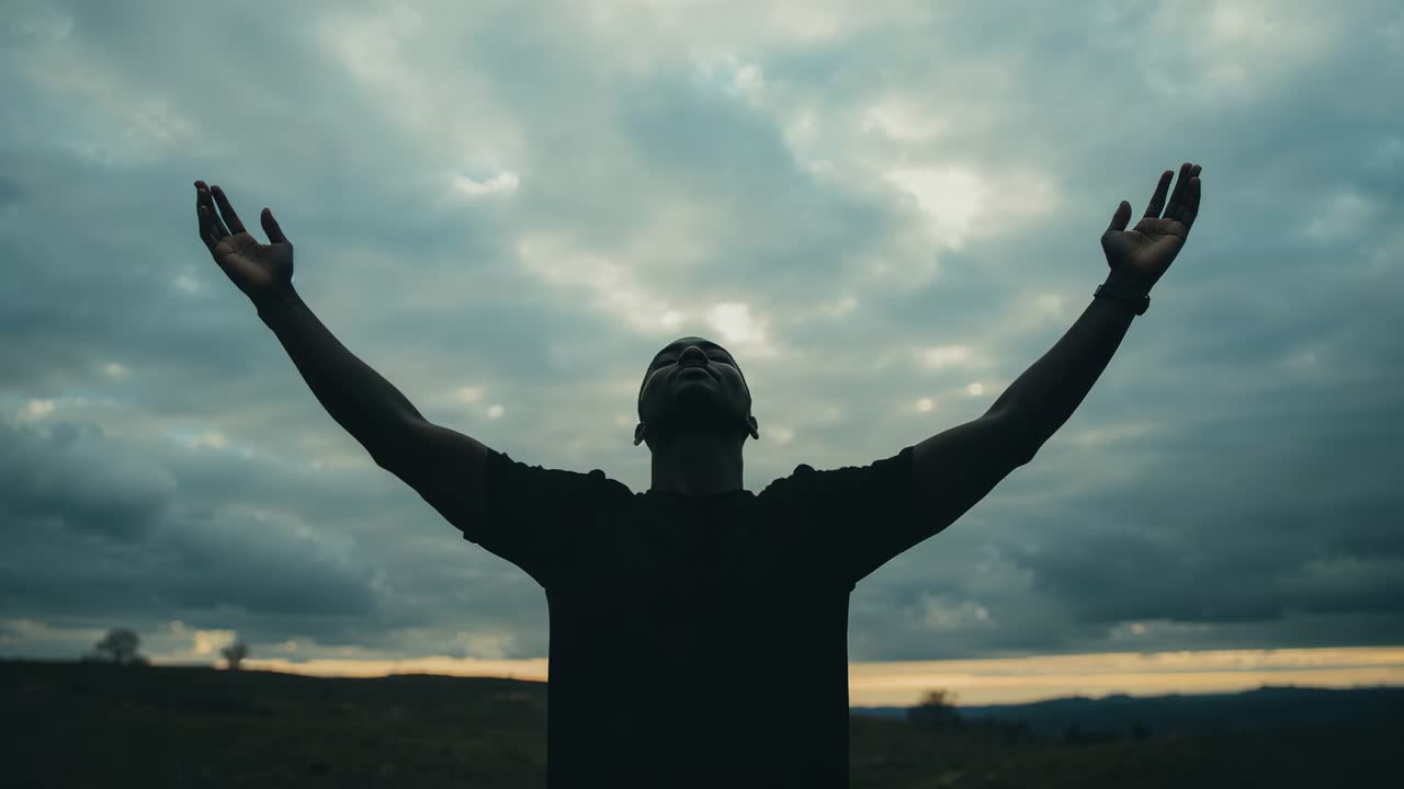 Reacting to sunrise light, raising man wearing dark shirt lifting arms toward cloudy horizon, watch