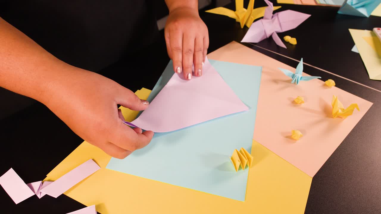 Primo piano dall'alto di mani che piegano con precisione carta viola per origami su un'area di lavoro vivace