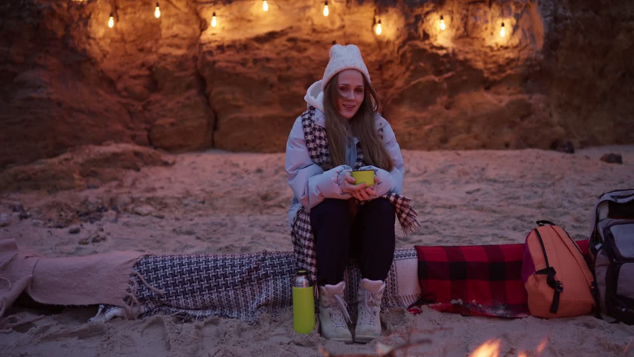 Woman enjoying a warm drink by a campfire on the beach at sunset