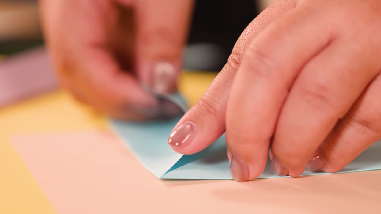 Nærbillede af hænder, der omhyggeligt folder lyseblåt papir på et skrivebord til origami-kunst