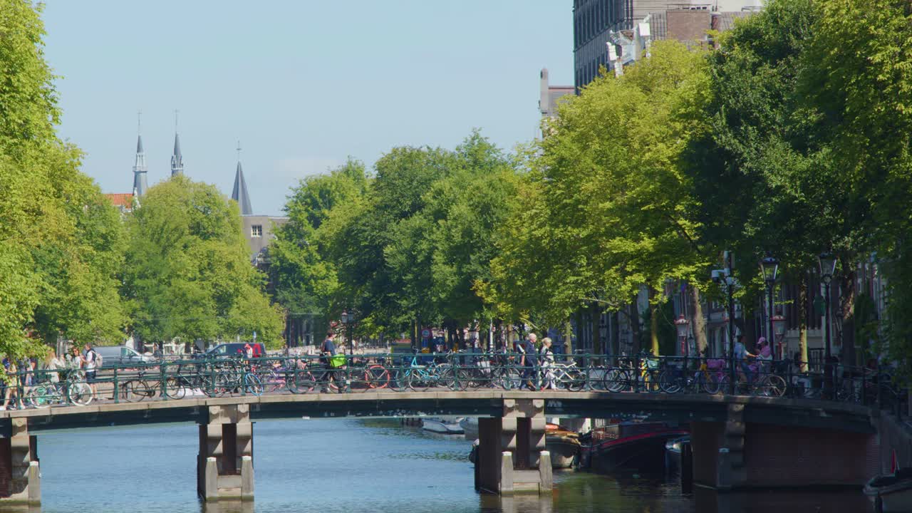 Bicyclists travel across a tree-lined bridge over canal, bright daylight, static wide shot