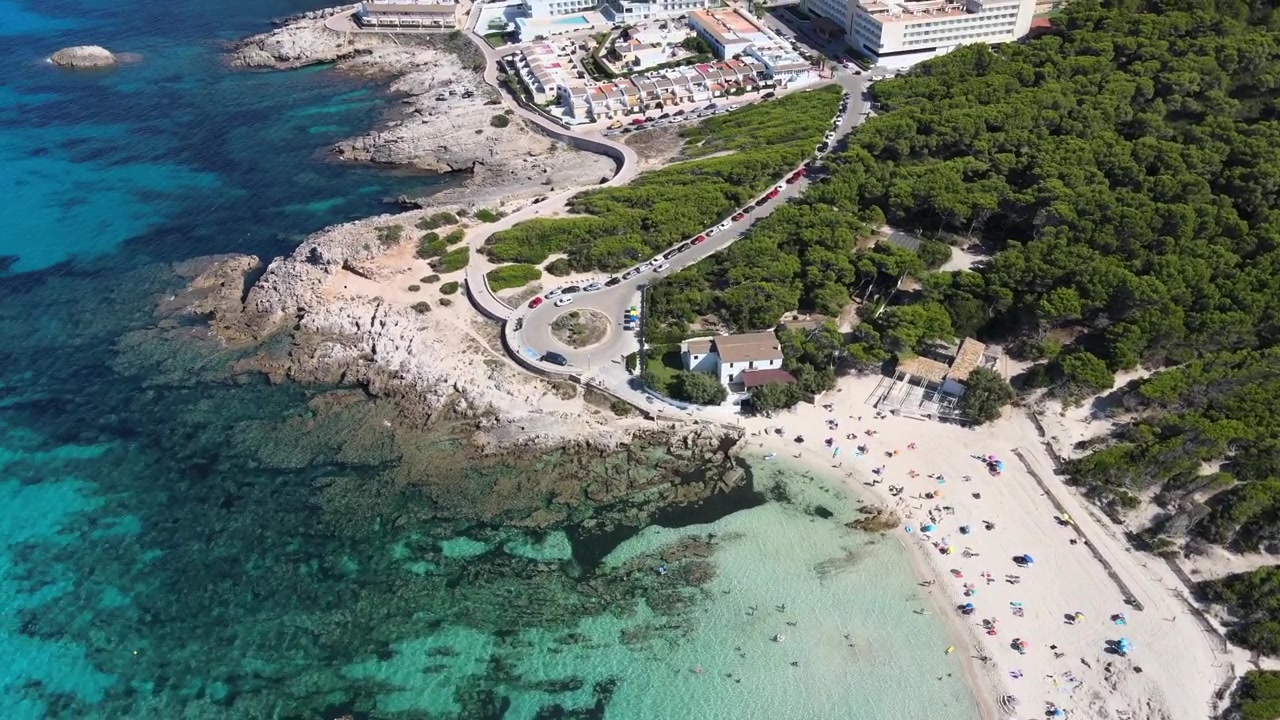 Banhistas desfrutam das areias macias e águas cristalinas de Cala Agulla em Cala Rajada, Maiorca. A vegetação exuberante proporciona um cenário deslumbrante para este popular destino de praia.