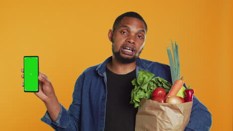 Young-guy-with-a-bag-of-fresh-groceries-showing-greenscreen-on-his-smartphone