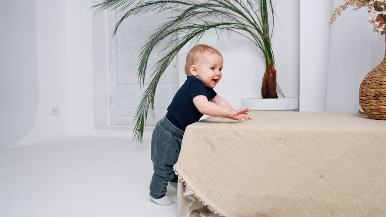 Cheerful baby boy in t-shirt and jeans stands at the table. Happy little child is in the room smiling and tapping by the surface.