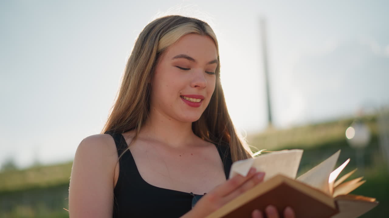 primer plano de una joven vestida de negro hojeando un libro al aire libre bajo un cielo brillante, encontrando papel escondido entre las páginas, mirándolo con curiosidad, sonriendo suavemente mientras la luz del sol ilumina suavemente su rostro
