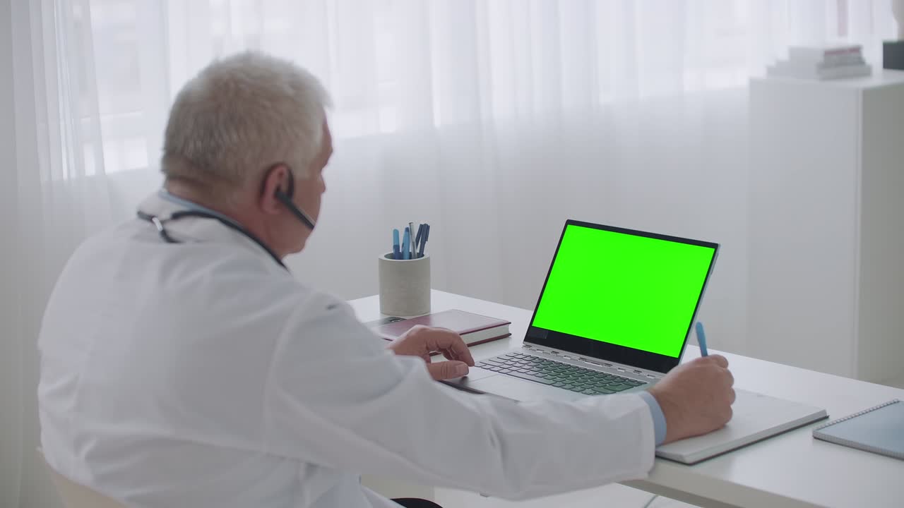 un médico de edad avanzada está escuchando un webinar o un paciente en una computadora portátil con pantalla verde para la tecnología chroma key, escribiendo notas en un cuaderno