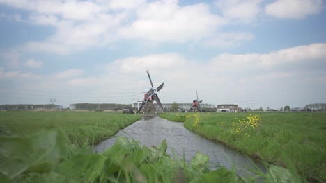 Windmills-In-The-Farm-With-Green-Grass-And-Stream-In-Foreground-In-Leiderdorp,-Leiden,-Netherlands