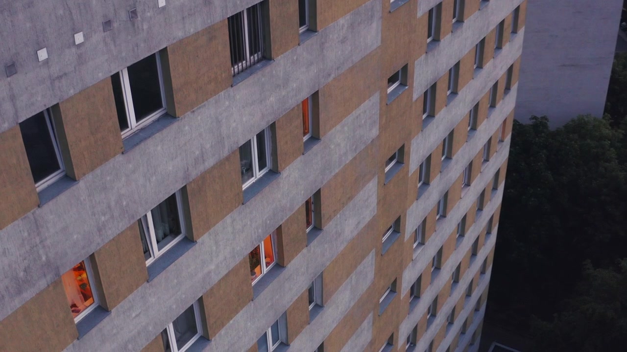 Toma abstracta de dron orbitando la fachada de un edificio de apartamentos de varios pisos color beige, de Europa del Este, con filas geométricas de ventanas, algunas iluminadas con luz cálida. Atardecer, atmosférico, cinematográfico
