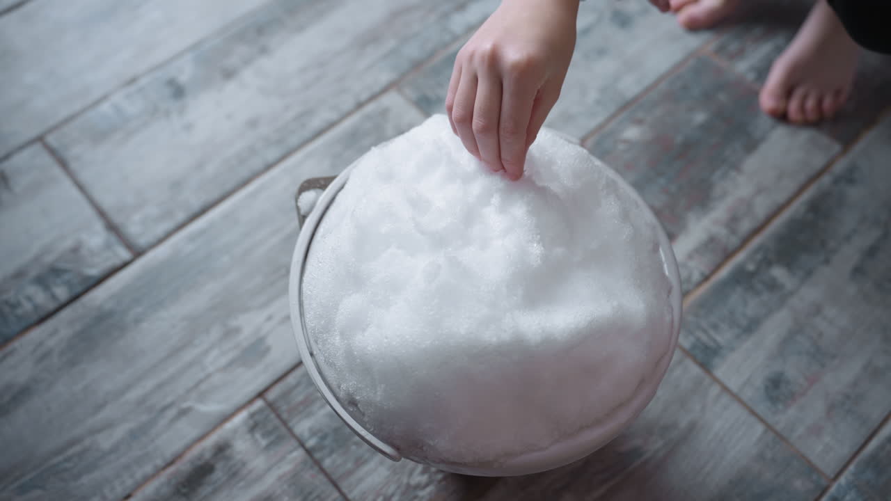Female hands dipping into container of melting snow on rustic tiled floor, fingertips pressing powdery mound, glistening droplets at edges, bare toes peeking in background under soft indoor light