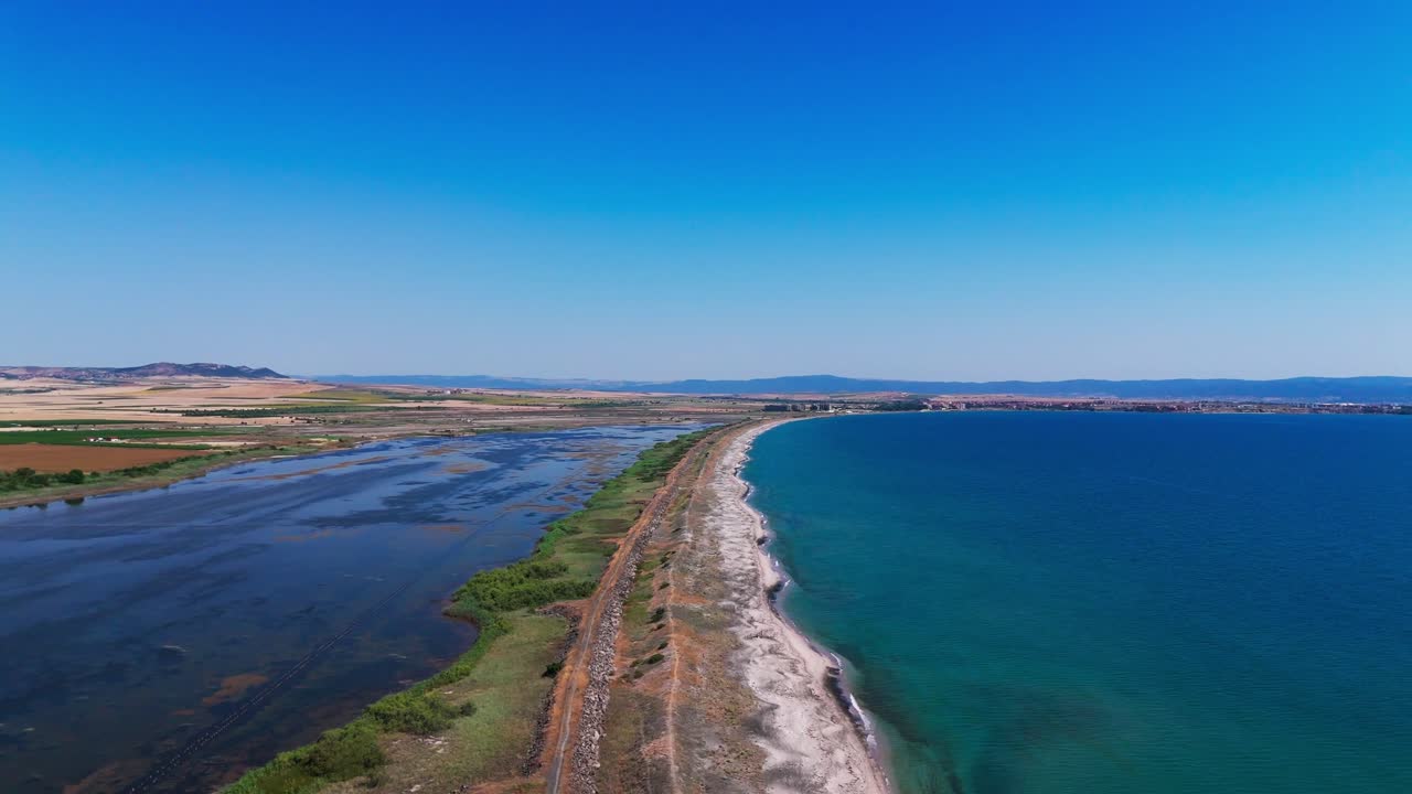 Un drone sorvola una stretta striscia di terra che separa il mare aperto dalle acque interne, con campi rurali sotto cieli sereni.