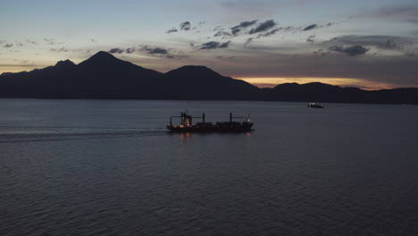 Cargo-Freight-Ship-Motors-Out-at-Night-through-Ocean-Channel-with-Mountains