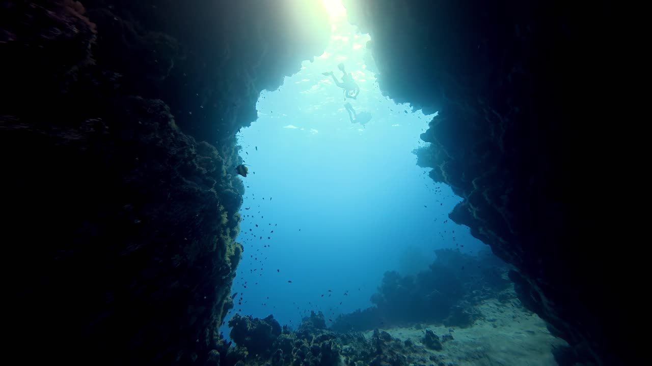 A luz do sol penetra a água azul cristalina em uma caverna subaquática escura durante o verão, enquanto mergulhadores silhuetados exploram o vibrante fundo de recife de coral, capturado de um ângulo baixo e amplo para cima.