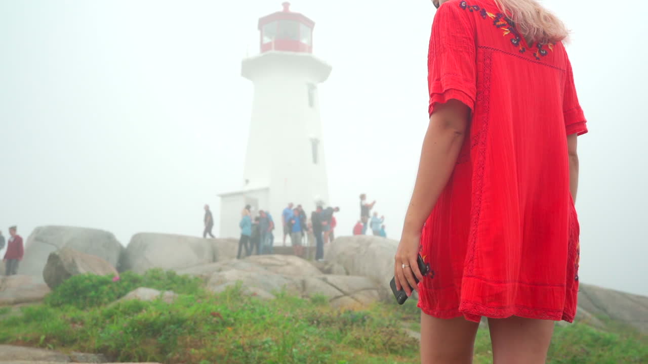 Jovem garota caminhando em direção a um farol branco na neblina em um dia nublado na Nova Escócia, Canadá, usando um vestido vermelho