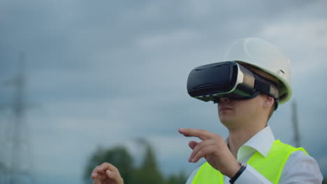 a male electrician in virtual reality glasses moves his hand simulating the work with the graphical interface of the power plant against the background of high-voltage electric transmission lines.