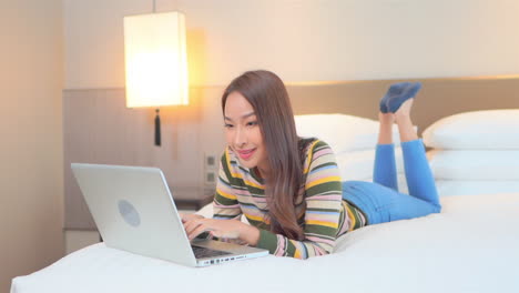 slow-motion of relaxed asian woman using her laptop computer lying on the bed with her legs raised at hotel room wearing casual clothes, working from home