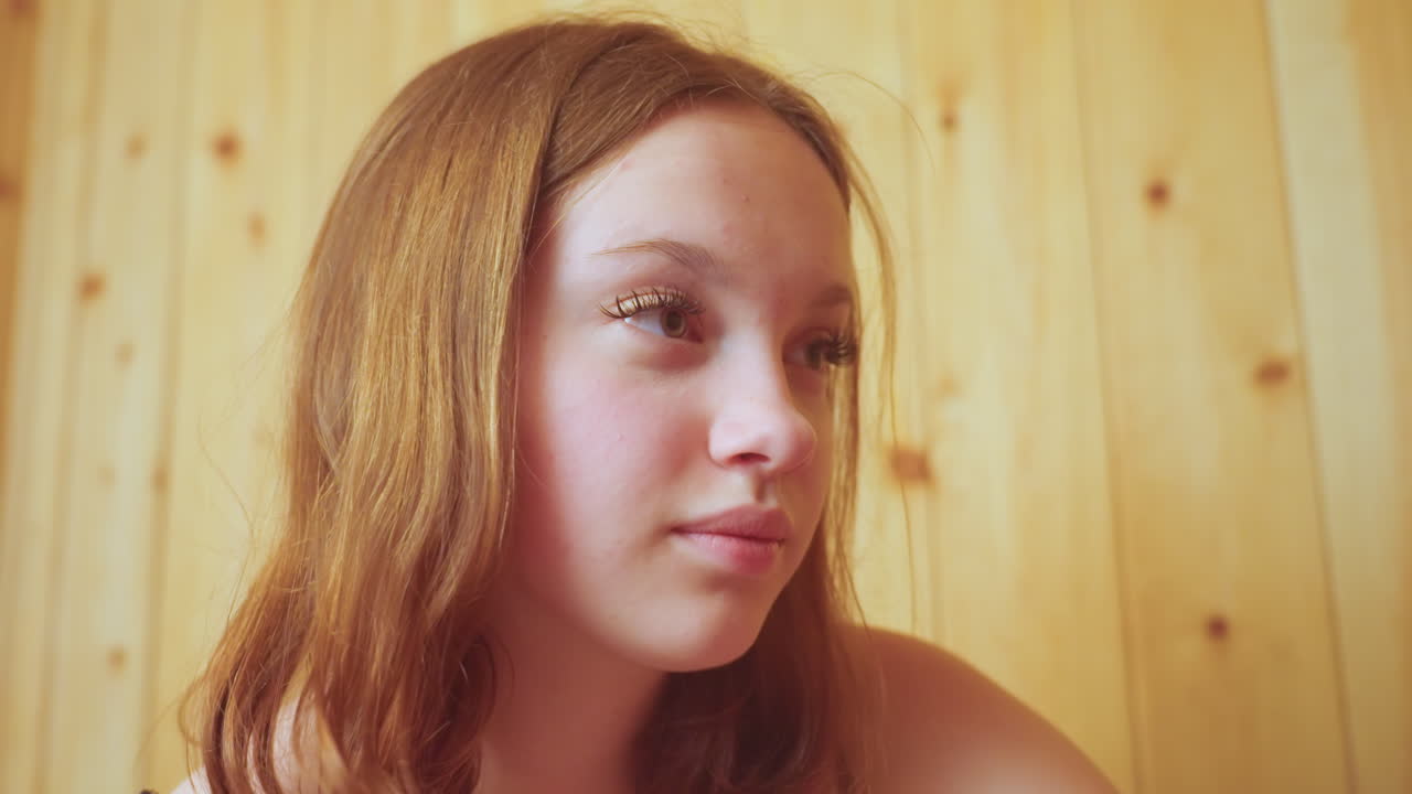 close up portrait of teenage girl looking thoughtful against wooden sauna wall soft warm light highlighting long lashes and serene expression calm reflective mood in cozy wellness setting