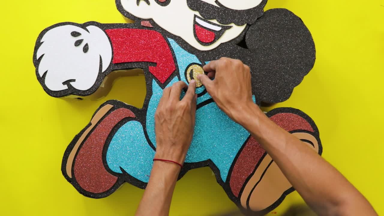 Hands assemble a video game character figure made of glittery foam pieces on a yellow background