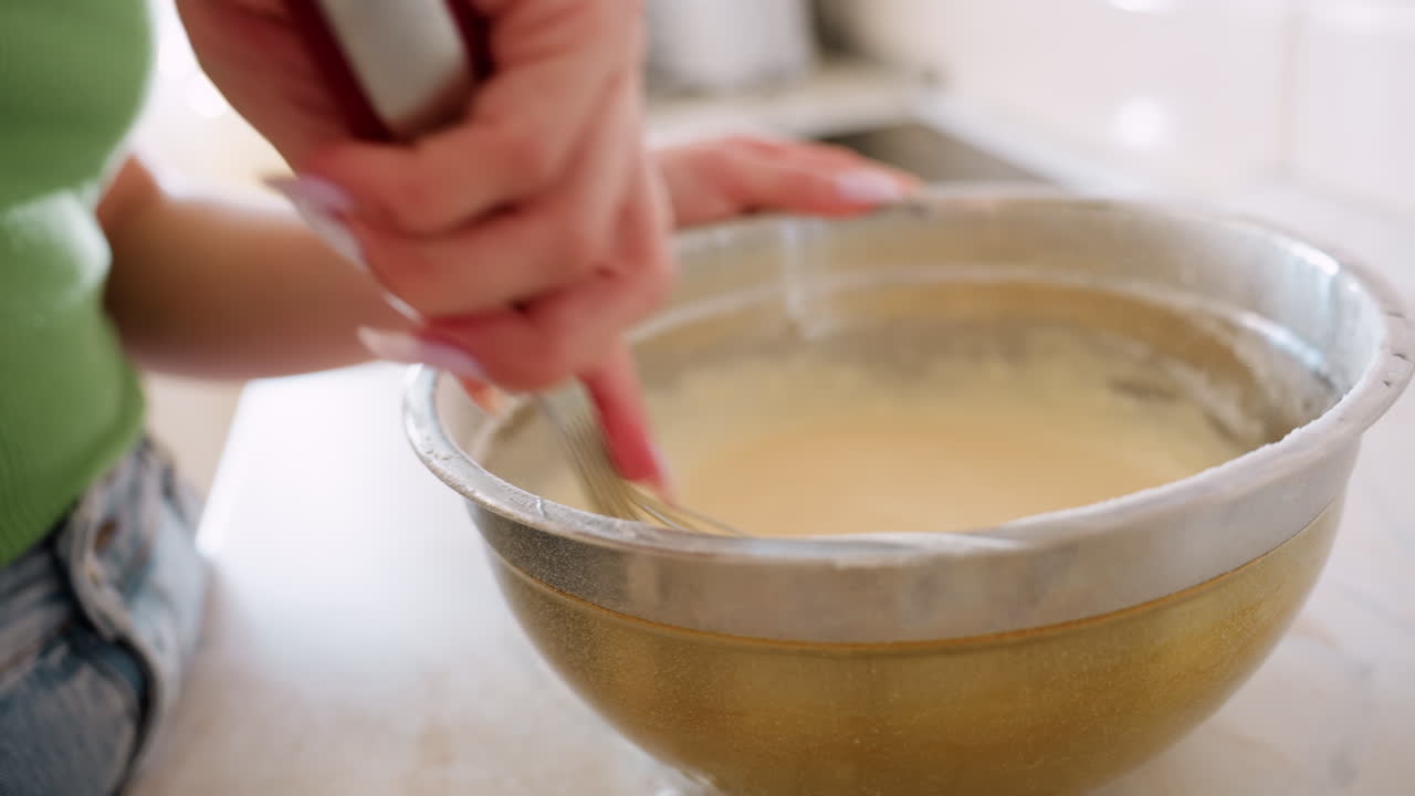 Cook preparing breakfast while stirring smooth batter in stainless bowl with whisk, focusing on close up hand movement as ingredients blend together during baking process in bright home kitchen
