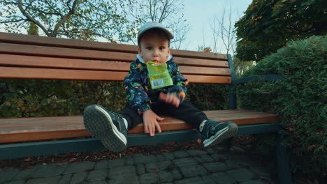 caucasian toddler in warm clothes sits on the bench in park. kid holding a pack of juice in mouth and tapping his hands by the bench. low angle view.