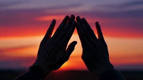 man creates a triangle shape with his hands, silhouetted against a stunning sunset filled with vibrant colors