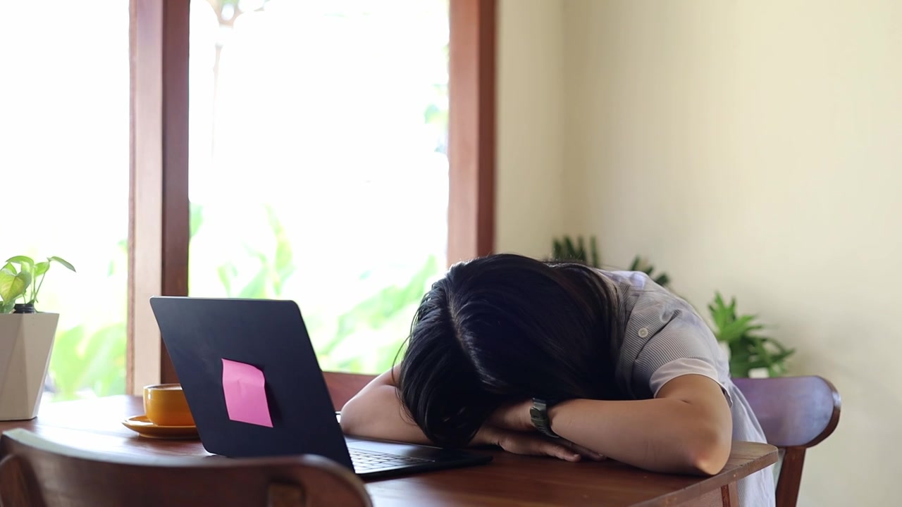 Ung asiatisk kvinne sover på skrivebordet på grunn av overarbeid og utmattelse mens hun jobber på bærbar PC på kafé.