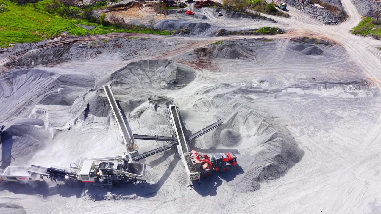 Close up elevated view of mechanical conveyor sorting aggregate piles with red wheel loader. Mobile rock crushing machine operating at industrial site