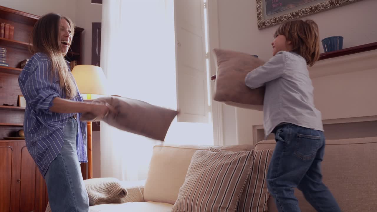 Glad kaukasisk familie, en ung mor og hendes lille søn, der hygger sig med en pudekamp på sofaen derhjemme.