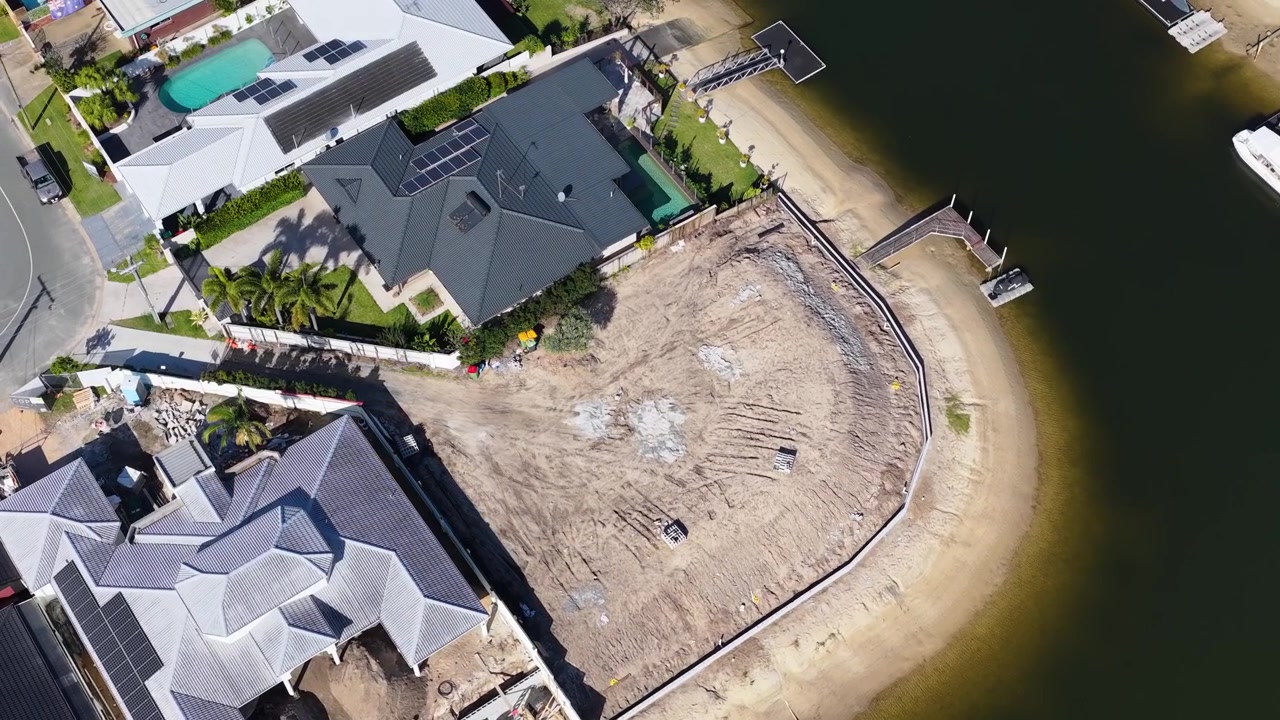 High-angle drone shot reveals residential construction progress beside luxury canal homes in bright daylight.
