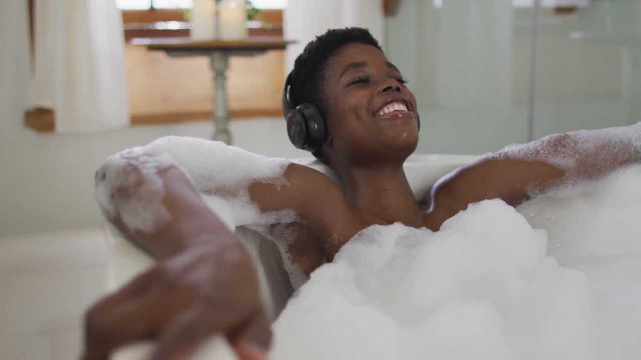 Smiling african american attractive woman wearing headphones relaxing in foam bath in bathroom