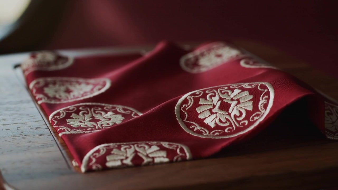 Shifting viewpoint nudging red textile flattening on wood tray to reveal embroidered medallions