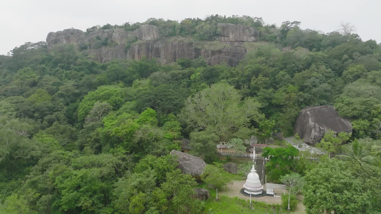 An aerial view of a serene Buddhist temple with a white stupa nestled at the base of a massive rock formation, surrounded by lush tropical forest in the heart of Sri Lanka