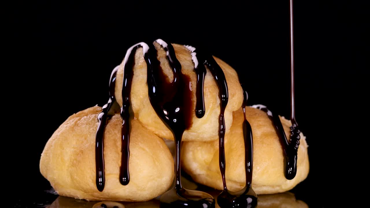 Three cream-filled choux pastries are stacked and slowly drizzled with glossy chocolate sauce against a black background, captured in close-up with dramatic lighting
